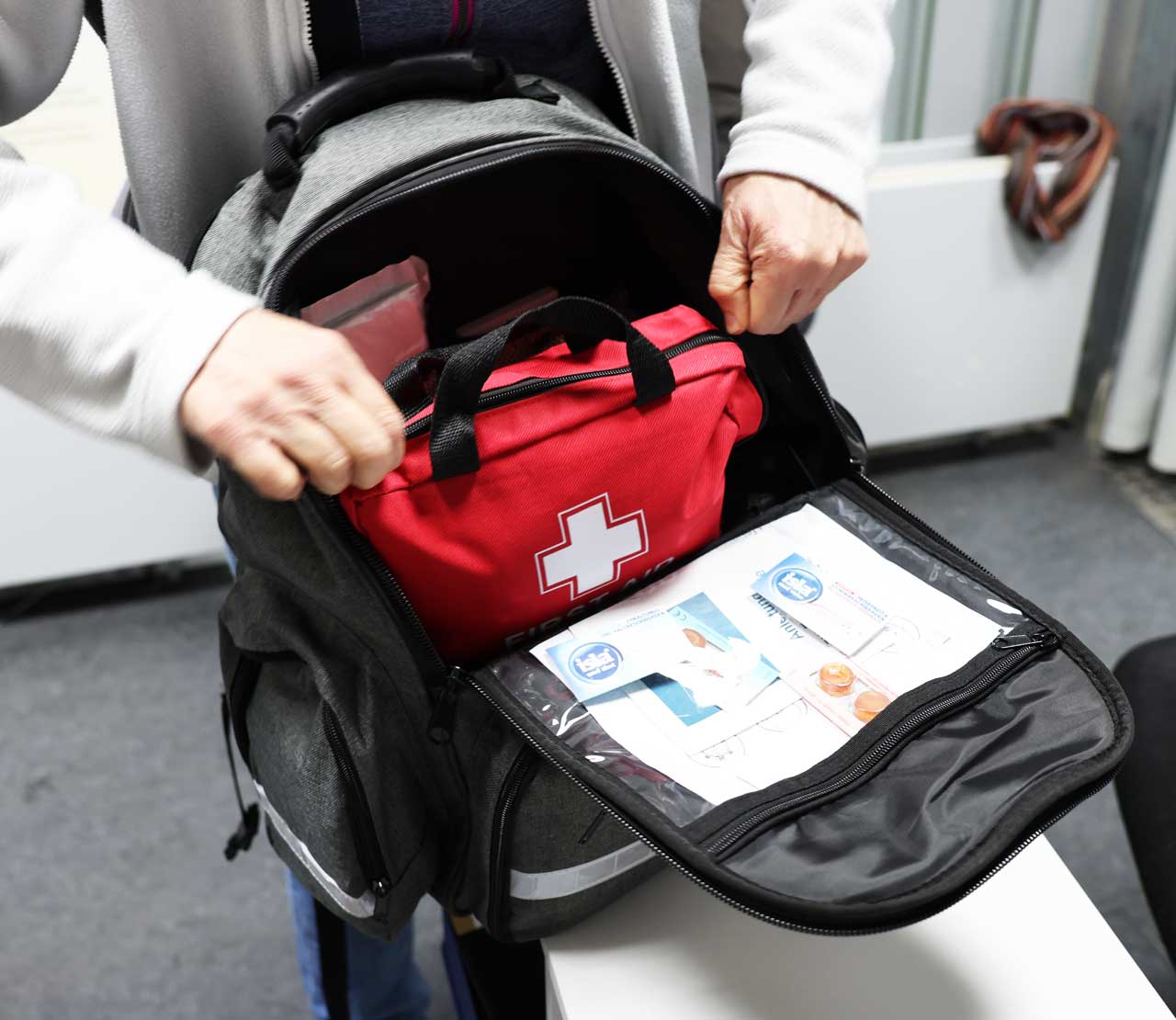 Geöffneter Rucksack, man sieht nur Hände und eine rote Erste-Hilfe-Tasche. 