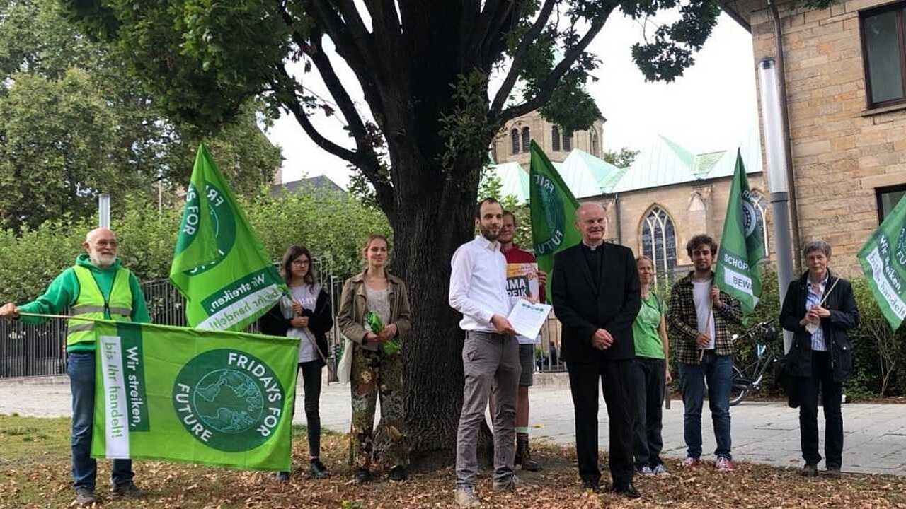 Mehrere Klimaaktivisten stehen unter einem Baum und übergeben Forderungen an Bischoff Overbeck.
