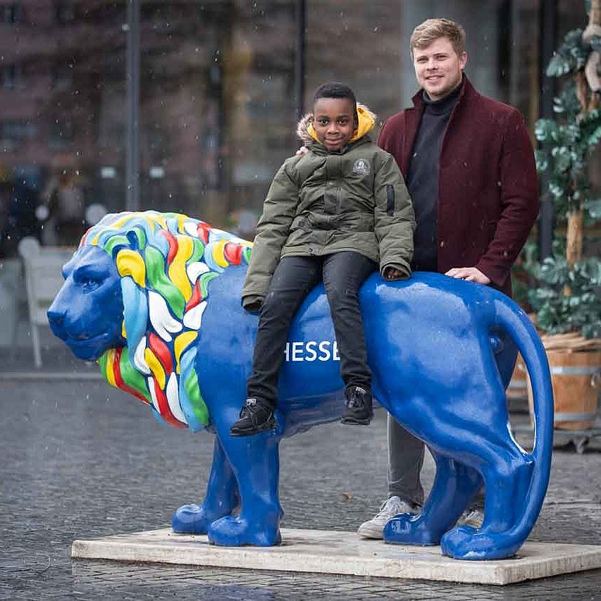 Richmond sitzt auf einem künstlichen blauen Löwen vor dem Eingang des Frankfurter Zoos. Mathias steht hinter ihm.