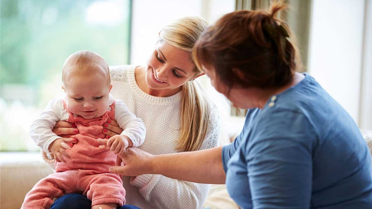 Eine junge Mutter mit einem Baby auf dem Schoß auf einer Couch. Davor sitzt eine ältere Person, sie streckt die Hand zum Baby hin, das Baby greift ihren Finger.