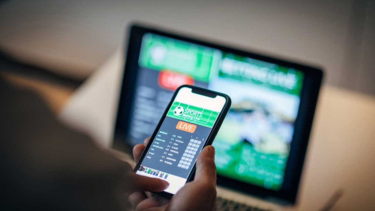 Eine Person hält ein Smartphone mit einer Live-Sportwetten-App in der Hand. Im Hintergrund ist ein unscharfer Laptop mit einer geöffneten Sportwetten-Website zu erkennen.