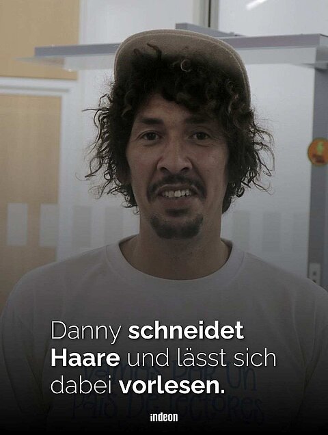 Zu sehen ist ein Foto von Danny Beuerbach. Er ist ein junger Mann, trägt eine braune Cap. Darunter stehen dunkelbraune Locken heraus. Er blickt direkt in die Kamera und trägt einen dunkelbraunen Oberlippen- und Kinnbart. Sein TShirt ist weiß. Darauf liegt ein dunkler Schatten. In weißer Schrift steht darauf: Danny schneidet Haare und lässt sich vorlesen. 