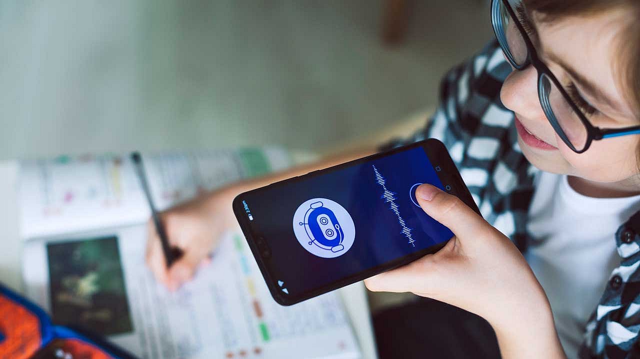 Ein Kind mit einer Brille hält ein Smartphone in der Hand vor seinen Mund. Auf dem Smartphone-Bildschirm ist eine App geöffnet, die ein stilisiertes Roboter-Symbol und eine Audiowellenform oder Soundwelle anzeigt. Es sieht aus wie eine Sprachassistenten-App oder eine KI-bezogene Anwendung.