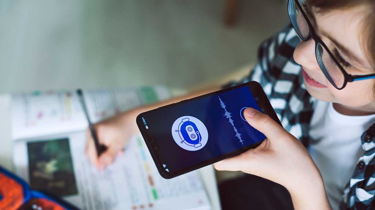 Ein Kind mit einer Brille hält ein Smartphone in der Hand vor seinen Mund. Auf dem Smartphone-Bildschirm ist eine App geöffnet, die ein stilisiertes Roboter-Symbol und eine Audiowellenform oder Soundwelle anzeigt. Es sieht aus wie eine Sprachassistenten-App oder eine KI-bezogene Anwendung.