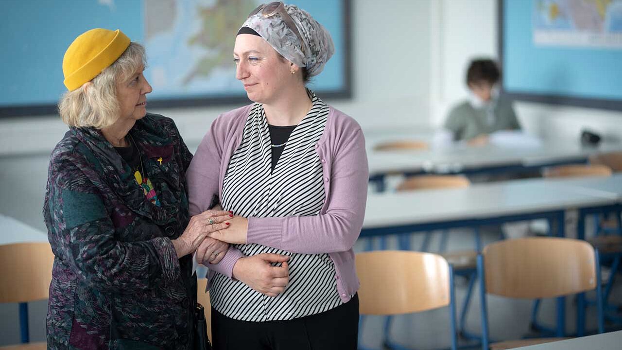 Die zwei Frauen stehen im Klassenraum Arm in Arm und schauen sich an. 