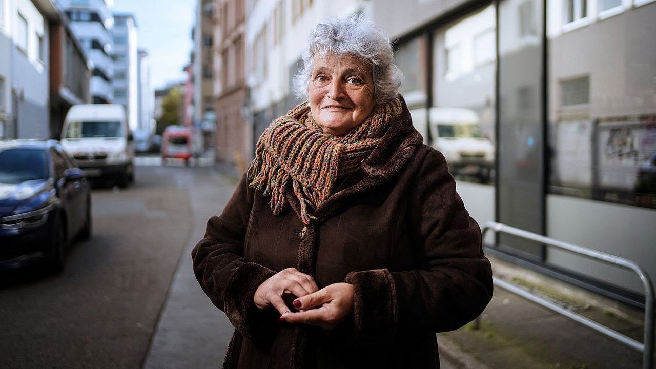 Eine ältere Dame mit grauen Haaren und dickem Mantel und Schal steht auf einer Straße, im Hintergrund sind Autos zu erkennen. Sie hat die Hände vor dem Bauch gefaltet und lächelt in die Kamera.