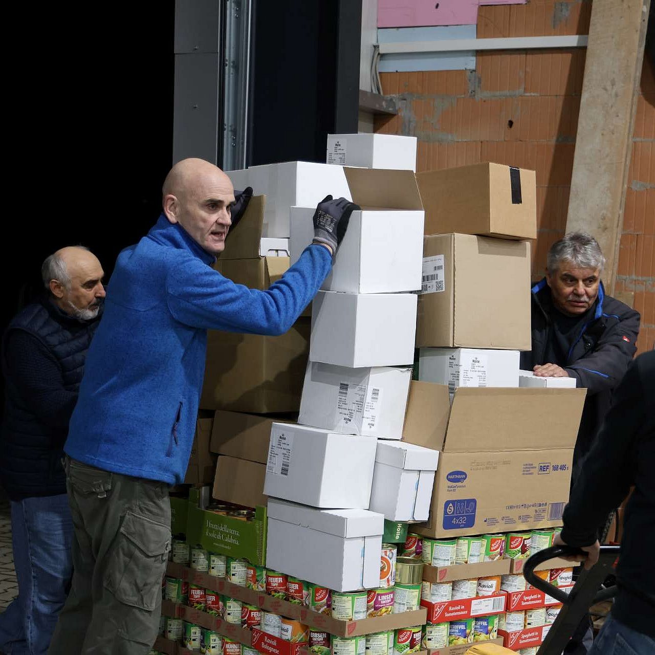 4 Ehrenamtliche fahren eine vollgepackte Europalette mit einem Hubwagen durch eine Lagerhalle