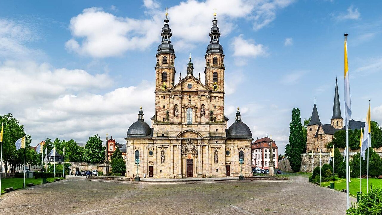 Das historische Domgebäude mit Domvorplatz. Rundherum Bäume und Fahnenmasten.