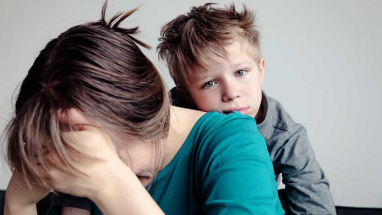 Frau beugt sich nach vorne und hat die Hände vor dem Gesicht. Hinter ihr ist ihr kleiner Sohn, der traurig in die Kamera schaut.