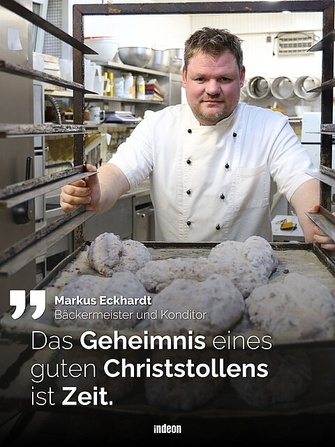Ein Bäcker in weißer Uniform steht in einer Bäckerei und hält ein großes Backblech mit Christstollen. Im Hintergrund sind Küchenutensilien zu sehen. Darunter steht der Text: „Das Geheimnis eines guten Christstollens ist Zeit.”