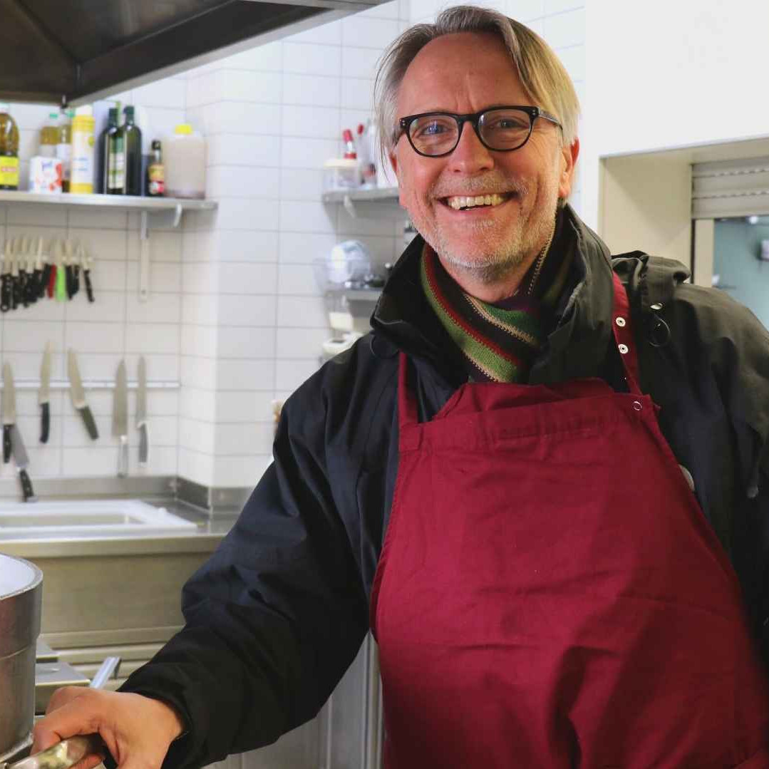 Thilo Schwarmann, ein Mitarbeiter der ada_kantine, steht neben einem großen Kochtopf und schaut lächelnd in die Kamera.
