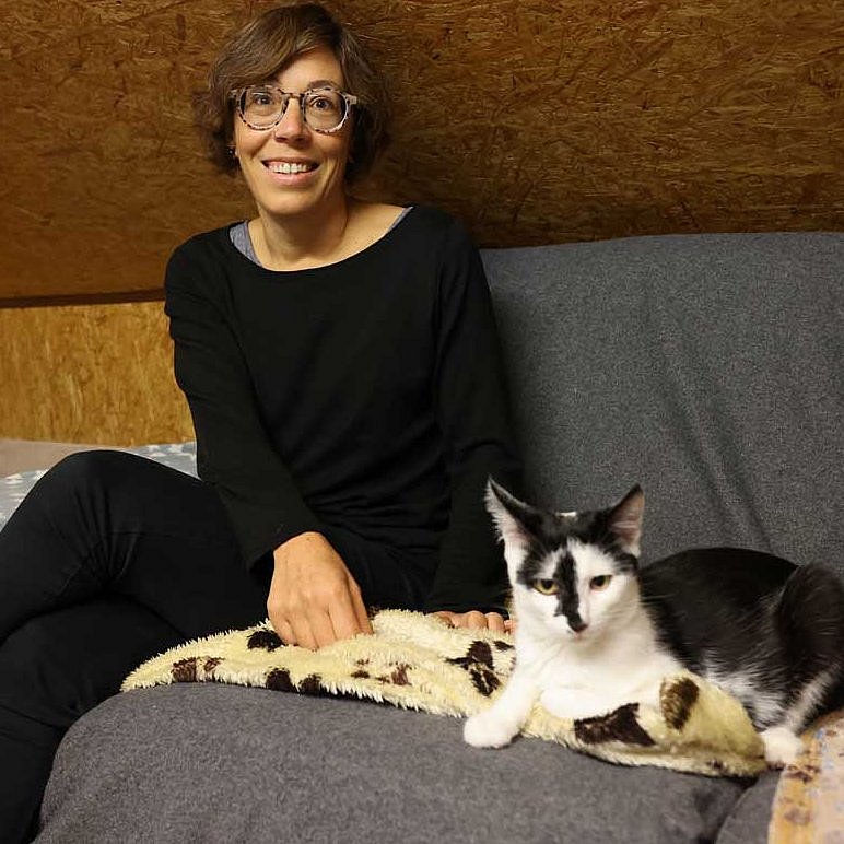 Marlene sitzt mit einer schwarz-weiß gestreiften Katze auf der Couch und lächelt in die Kamera.
