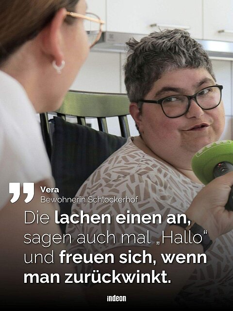 Eine Frau in einer Küche mit kurzen Haaren und Brille wird von einer zweiten Frau interviewt, sie hält ein grünes Mikrofon und trägt ein weißes Shirt. Text: Vera, Bewohnerin Schlockerhof: „Die lachen einen an, sagen auch mal Hallo und freuen sich, wenn man zurückwinkt."