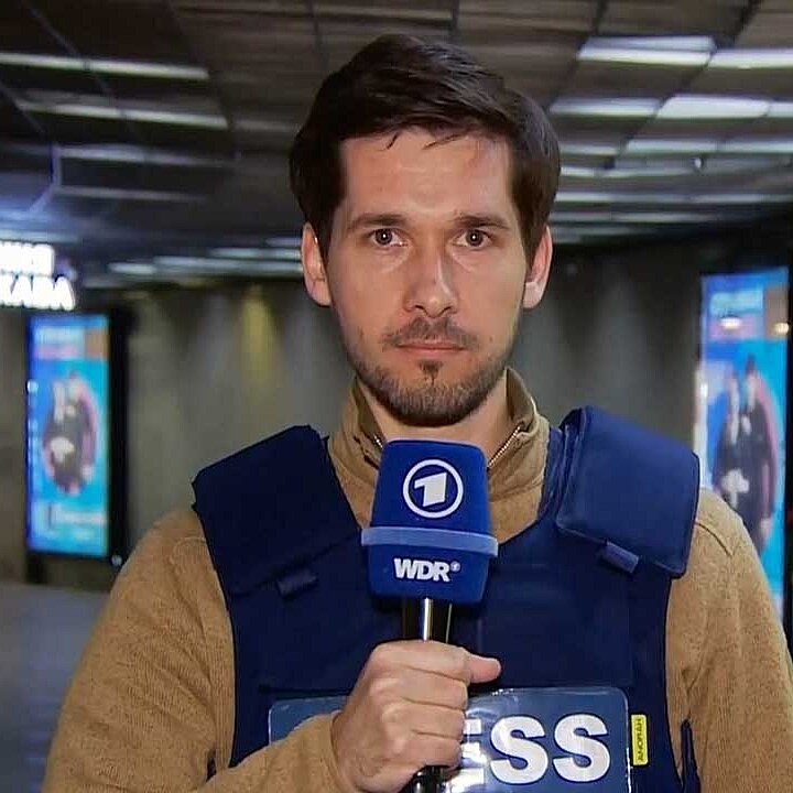ARD Reporter Vassili Golod während einer Liveschalte mit Schutzweste an und Mikrofon in der Hand 