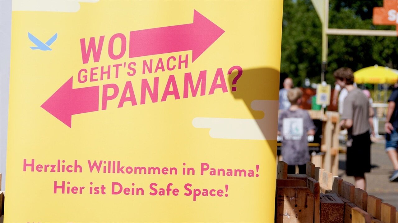 Ein gelbes Schild mit roter Schrift. Text: "Wo geht's nach Panama? Herzlich Willkommen in Panama! Hier ist dein Safe Space". Daneben mehrere Menschen unter Sonnenschirmen auf dem Festival