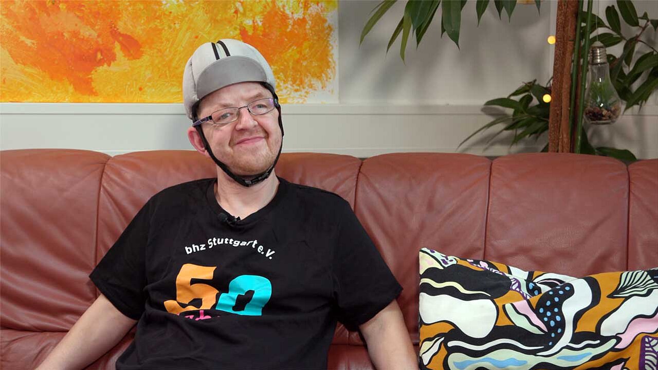 Christian sitzt auf einer rosa Leder-Couch. Er trägt eine Cappi, die unter dem Kinn verschlossen ist.