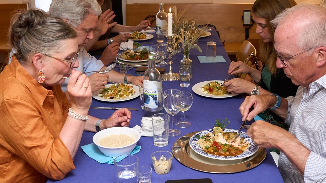 Mehrere Personen an einem gedeckten Tisch. Manche haben üppige Speisen vor sich, andere nur eine Schüssel Porridge. Auf dem Tisch stehen Wasserflaschen und Gläser, Kerzen und Tischdeko und Papierservietten in hellblau, die Tischdecke ist dunkelblau