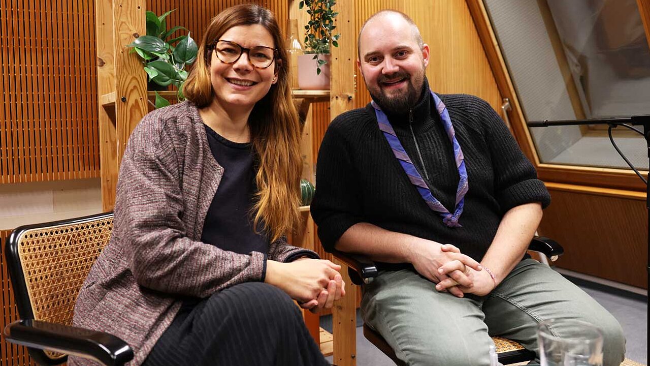 Charlotte und Jonas sitzen im gemütlichen Podcaststudio und lächeln in die Kamera.