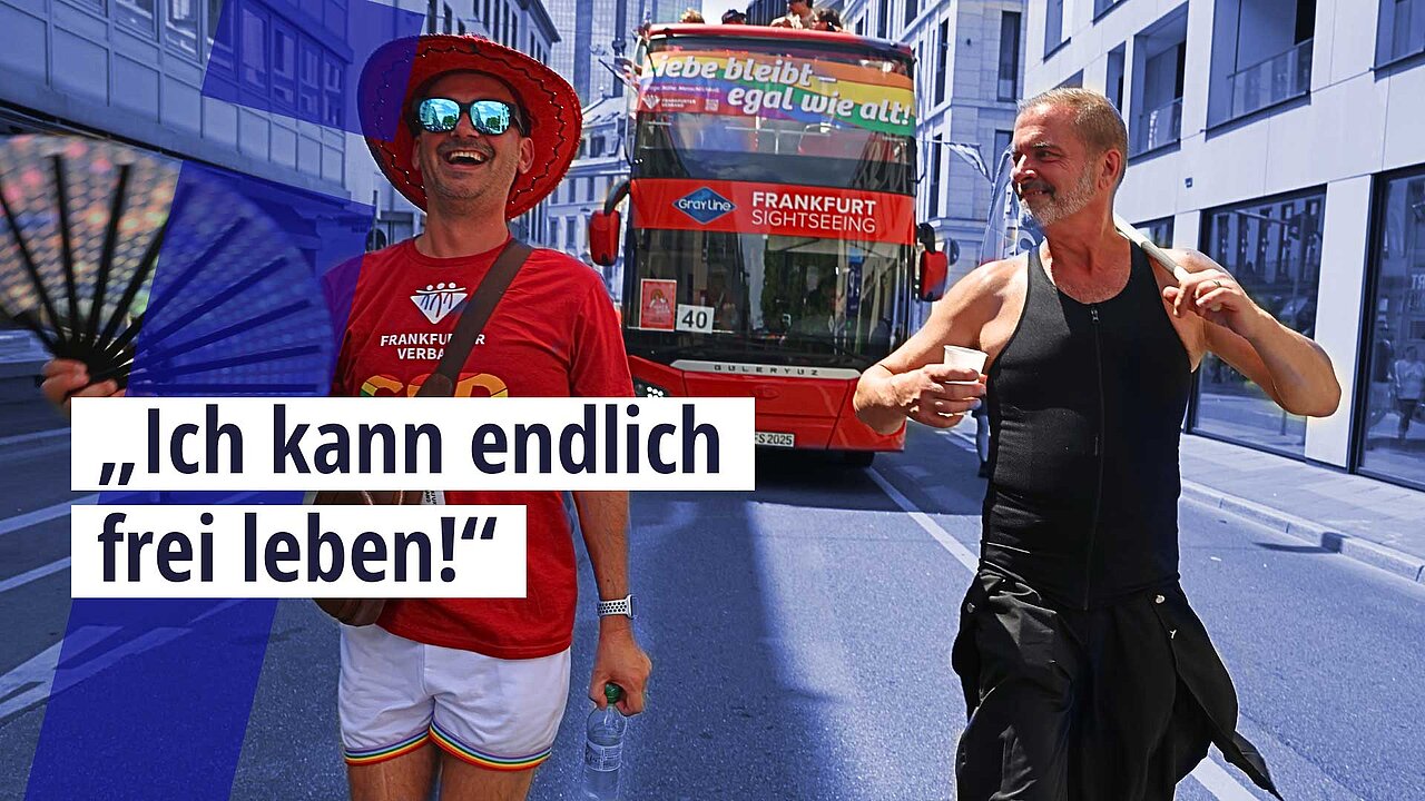 Thomas Kämmer und Armin Steinbauer gehen Seite an seite über die Straße. Es ist CSD, sie tragen Regenbogen Flaggen und Fächer. hinter ihnen fähnrt rin großer roter Dobbeldecker Bus mit Regenbogenbanner. 
