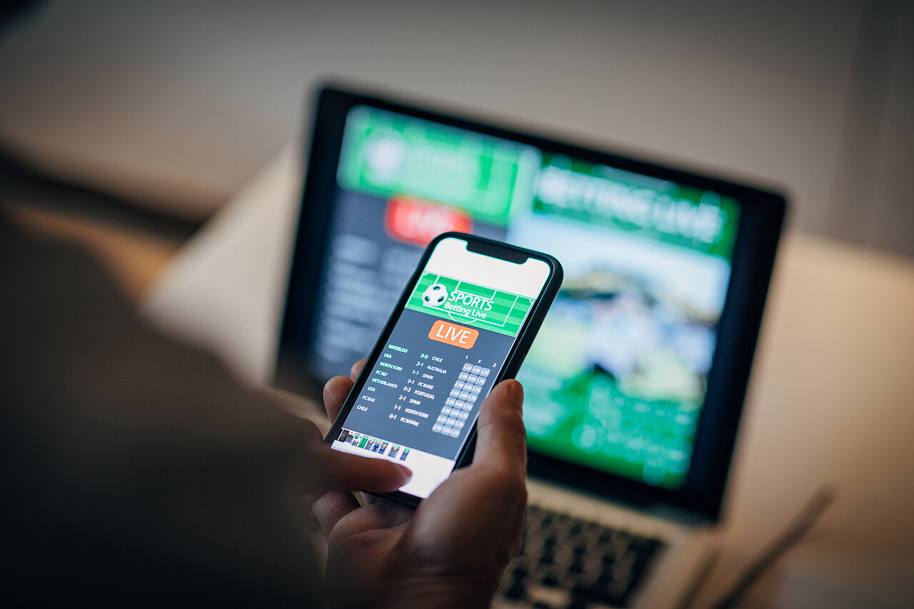 Eine Person hält ein Smartphone mit einer Live-Sportwetten-App in der Hand. Im Hintergrund ist ein unscharfer Laptop mit einer geöffneten Sportwetten-Website zu erkennen.