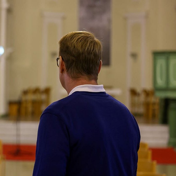 Mittelalter Mann mit Brille, weißem Hemd und blauen Pullover steht mit dem Rücken zur Kamera in einem Kirchenraum  