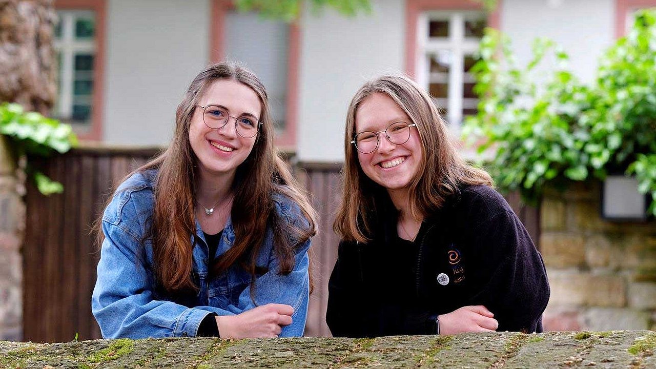Nina Maskus (rechts) und ihre Freundin Luisa Eizenhöfer stützen sich lächelnd an einer Mauer ab.