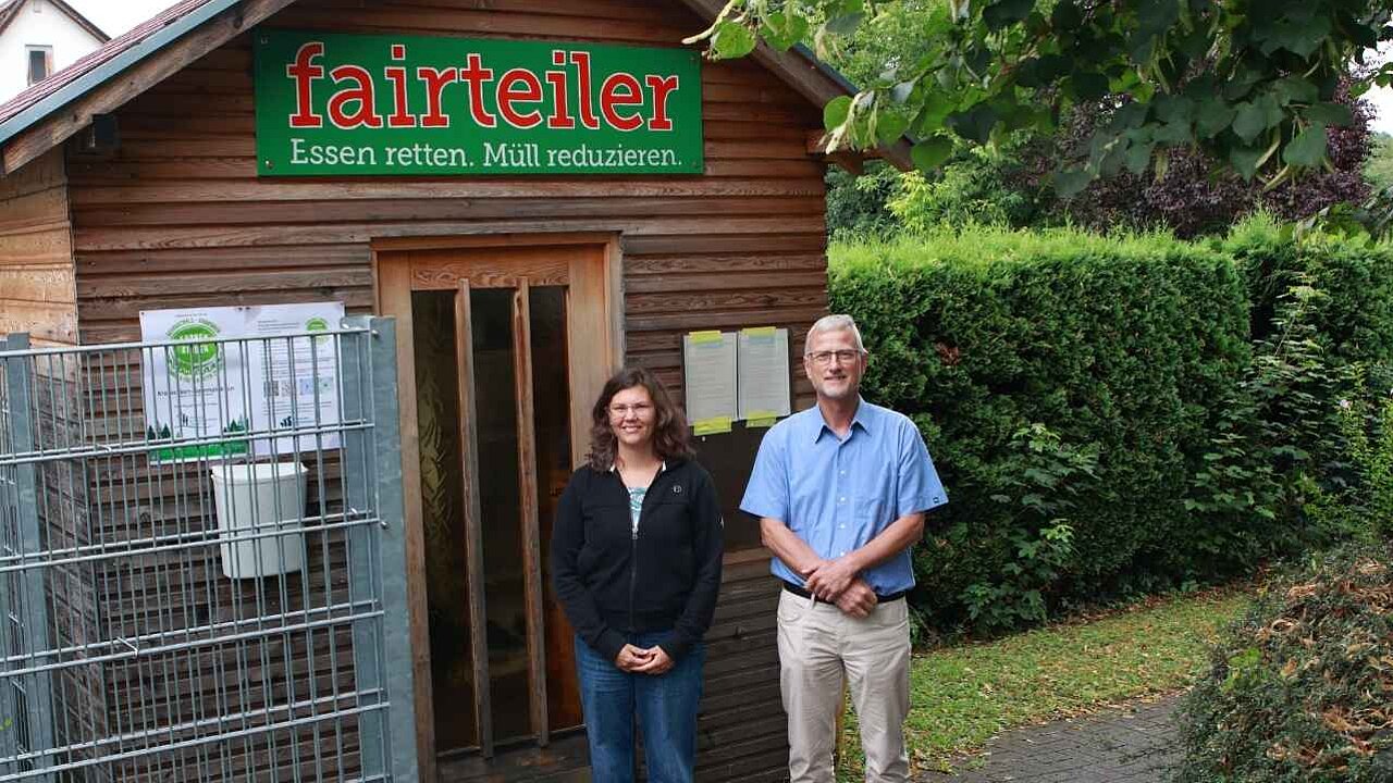 Eine 36-Jährige Frau mit dunkelbraunen langen Haaren und Pfarrer Markus Hägele, der eine hellblaues Hemd trägt, stehen vor dem Fairteiler in Scharnhausen. Das ist eine Holzhütte mit einem Schild darauf, in der sich Kühlschränke und eine Kommode befinden.