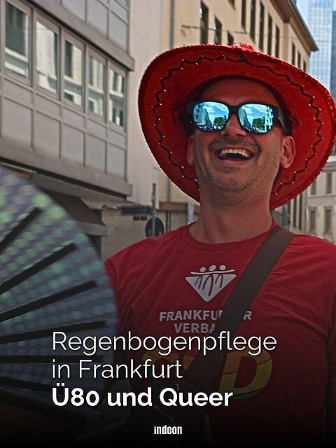 Ein Mann mit rotem Shirt, rotem Hut und verspiegelter Sonnenbrille auf dem CSD in Frankfurt. Er hält einen schillernden Fächer in der Hand. Text: Regenbogenpflege Frankfurt Ü80 und Queer