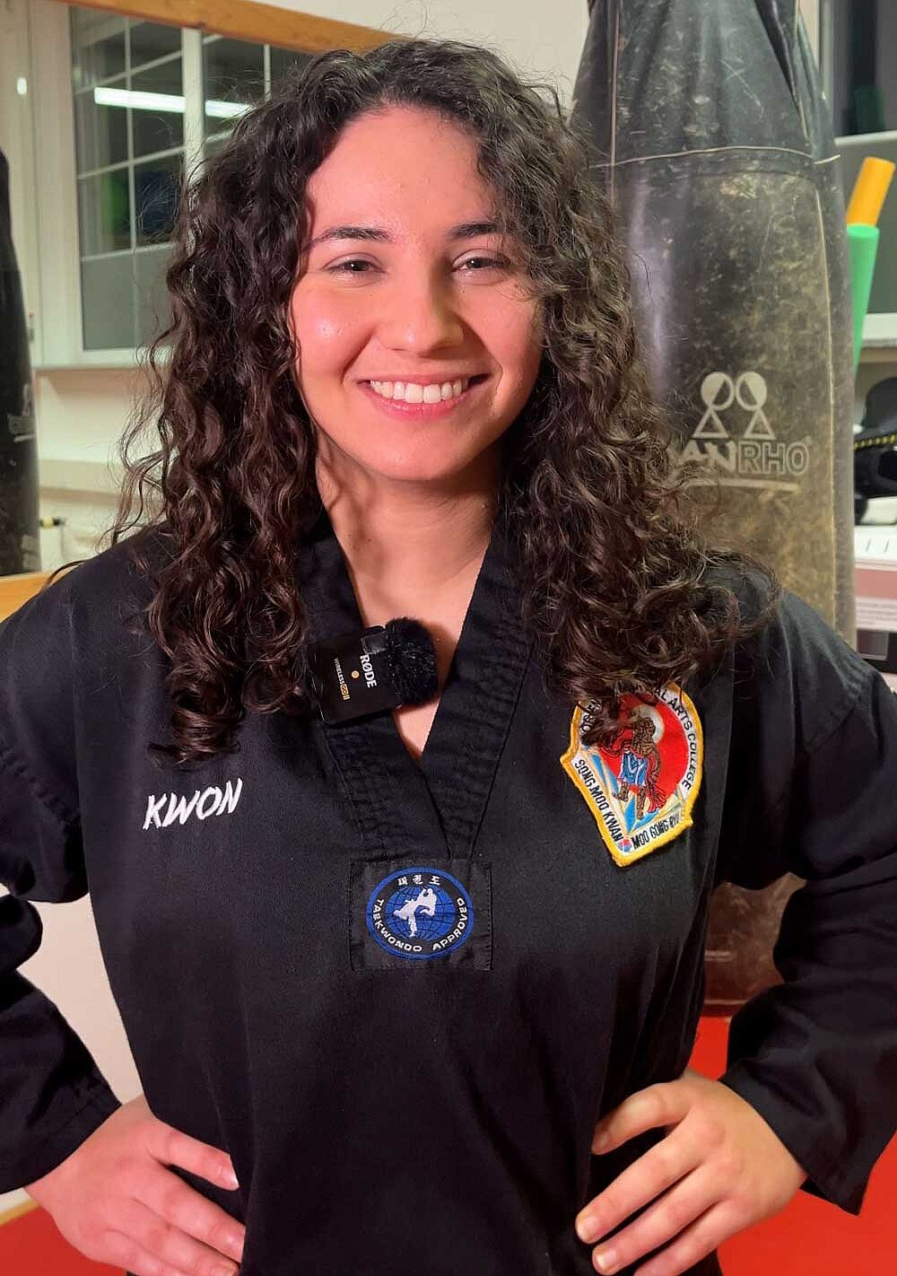 Aylin Çaka steht im Kampfsport-Gym und posiert mit den Händen ander Hüfte. Sie lächelt selbstbewusst und hat Locken. 