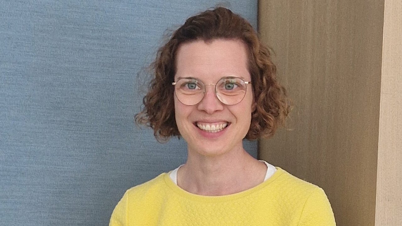Eine Frau mit braunen Locken, Brille und gelbem Pullover lächelt freundlich.