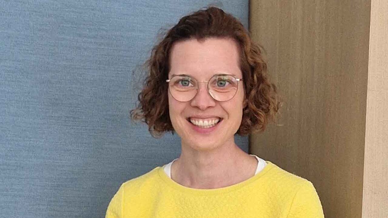 Eine Frau mit braunen Locken, Brille und gelbem Pullover lächelt freundlich.