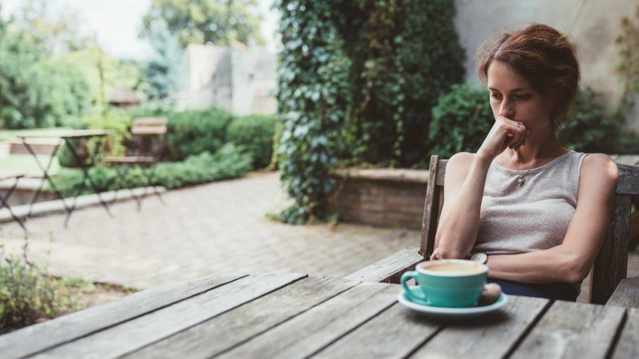 Eine vielleicht depressive Frau trinkt Kaffee auf der Veranda. Sie sieht unglücklich aus und hat eine Hand vor dem Mund, weil sie nachdenkt.