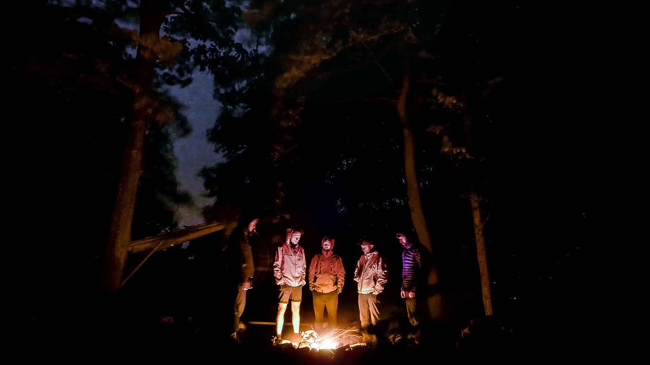 Dunkler Wald, Männer stehen um Lagerfeuer, in der Mitte ist es hell 