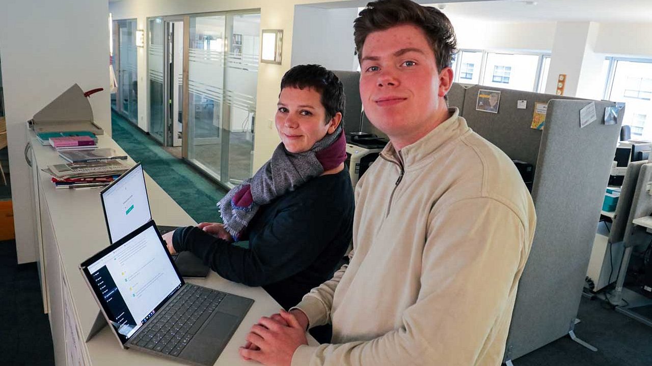 Esther und Christian stehen mit ihren Rechnern in der Mitte des Raumes. Sie blicken in die Kamera