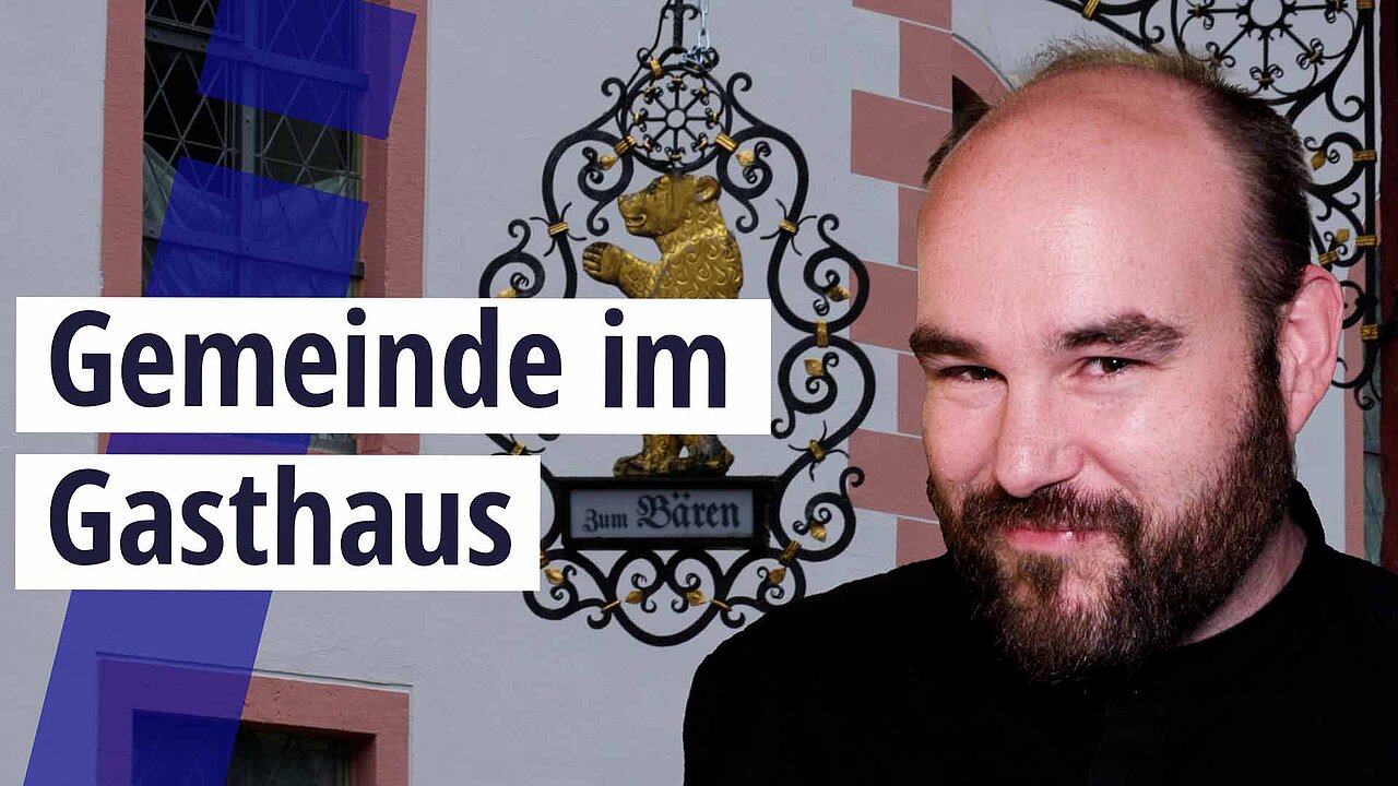 Eine historische Hausfassade mit rotem Sandstein und weißem Putz, daran hängt ein goldener Bär in einem schmiedeeisernen Schild. Im Vorderund ein Mann mit Halbglatze und dunklem Bart, er lacht freundlich. Text: Gemeinde im Gasthaus