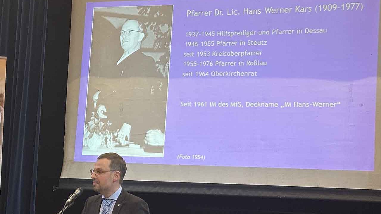 Historiker Jan Brademann spricht über die Kirche in der DDR an einem Pult. Im Hintergrund ein Foto (aus dem Jahr 1954) von Pfarrer Dr. Lic. Hans-Werner Kars (1909-1977). Dazu die Daten: 1937-1945 Hilfsprediger und Pfarrer in Dessau, 1946-1955 Pfarrer in Steutz, seit 1953 Kreisoberpfarrer, 1955-1976 Pfarrer in Roßlau, seit 1964 Oberkirchenrat, Seit 1961 IM des MfS, Deckname "IM Hans-Werner"