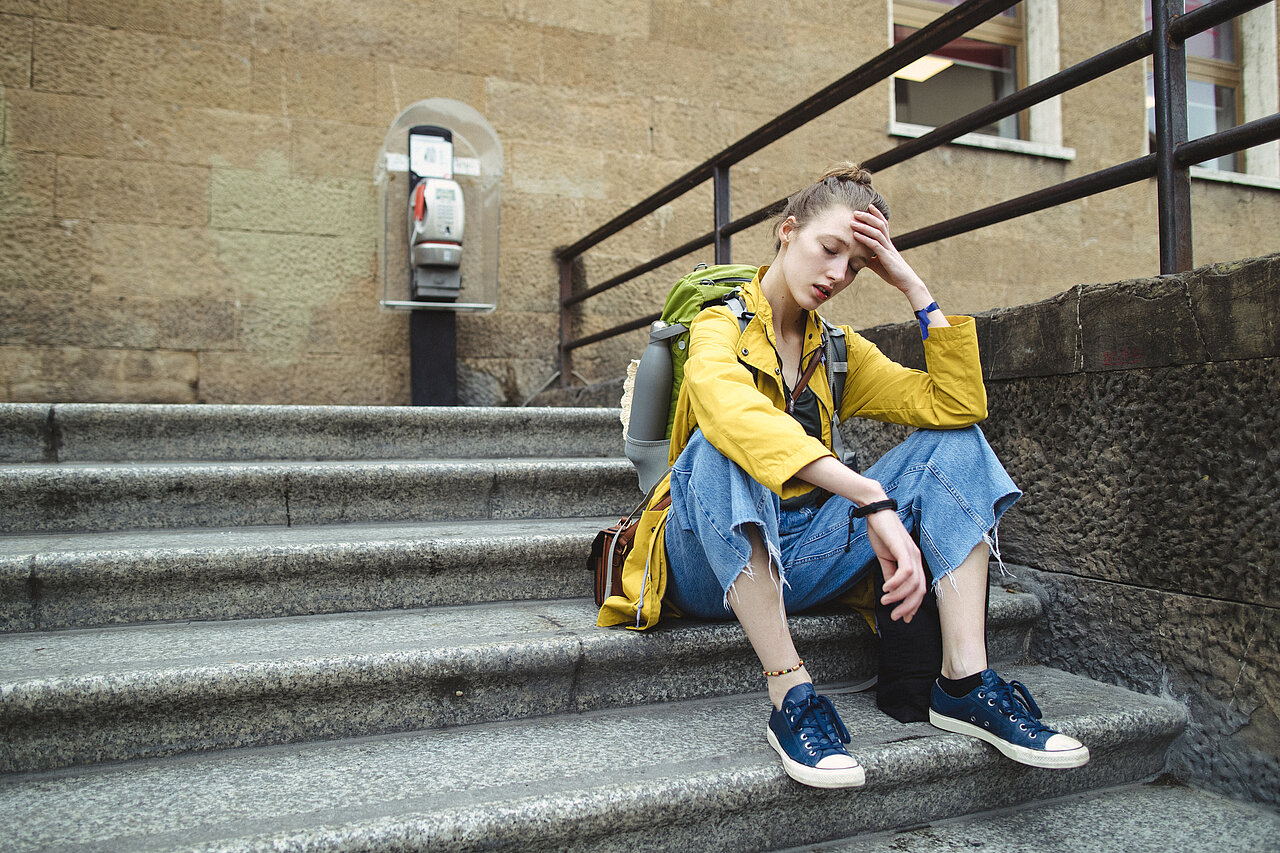 Das Bild zeigt eine junge Person, die mit Rucksack auf einer Steintreppe sitzt und sich erschöpft oder nachdenklich die Hand an die Stirn hält.