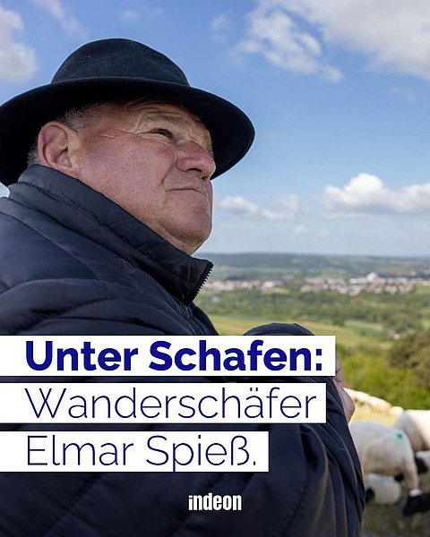 Wanderschäfer Elmar Spieß: ein weißer Mann, etwa 60 Jahre alt, mit schwarzem Hut und warmer Jacke sitzt auf einer Weide in der Rhön. Im Hintergrund der Ausblick über das Biosphärenreservat und einige seiner Rhönschafe