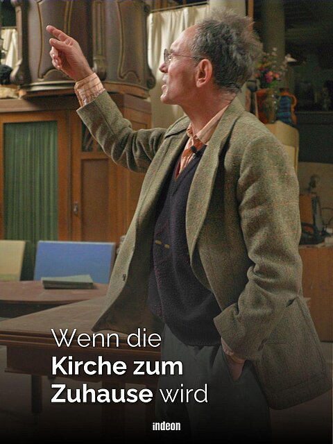 Ein älterer Mann im Sakko in einer alten Kirche. Im Hintergrund die Kanzel. Um ihn herum Tisch, Stühle und verschiedene Kunstwerke. Text: "Wenn die Kirche zum Zuhause wird"