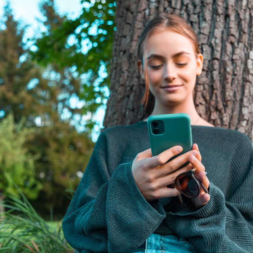 Junge Frau lehnt an einem Baum, lächelt selbstbewusst und blickt auf ihr Smartphone