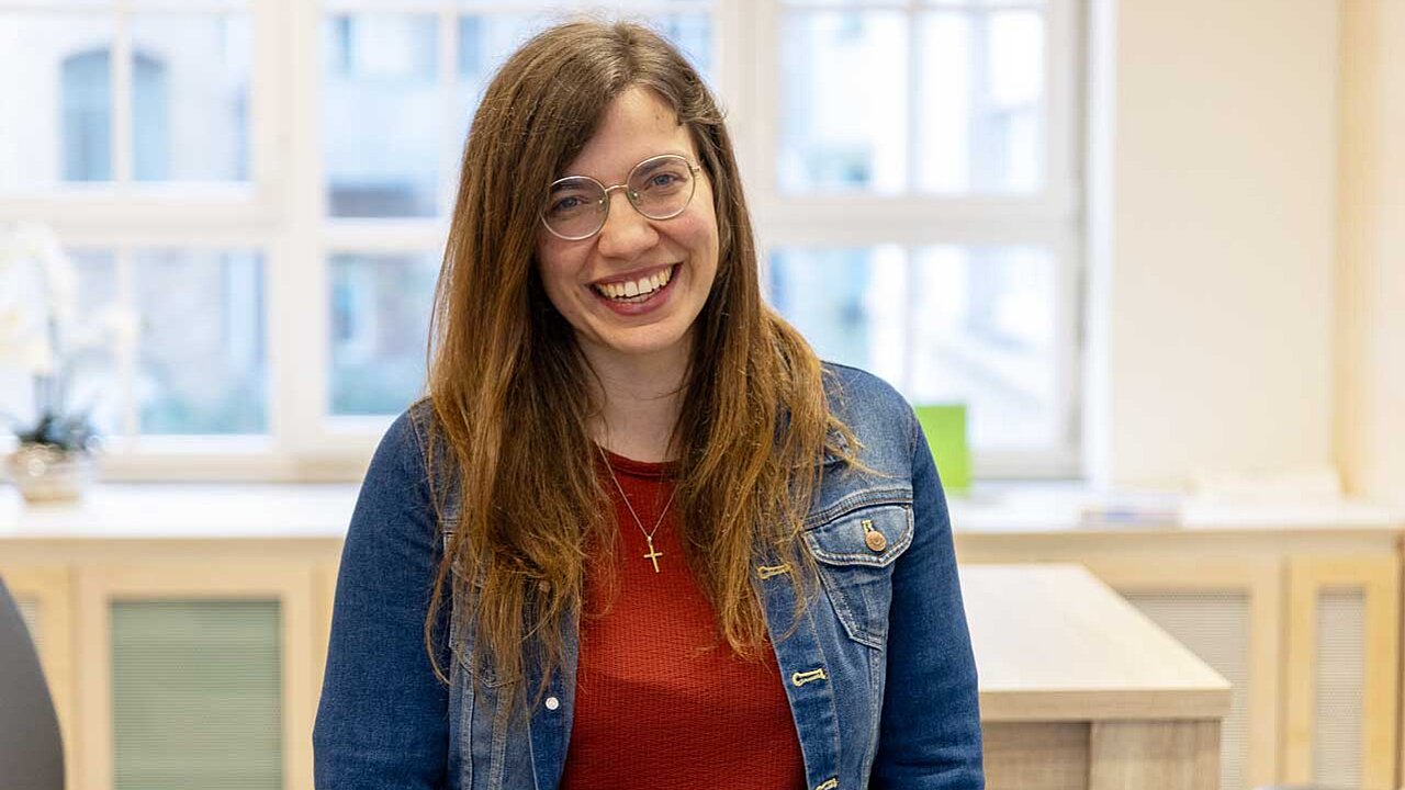 Ulrike steht in einem hellen Raum und lächelt in die Kamera. Sie hat lange Haare, trägt eine Brille und ein kleines Goldkreuz über ihrem roten Pulli.