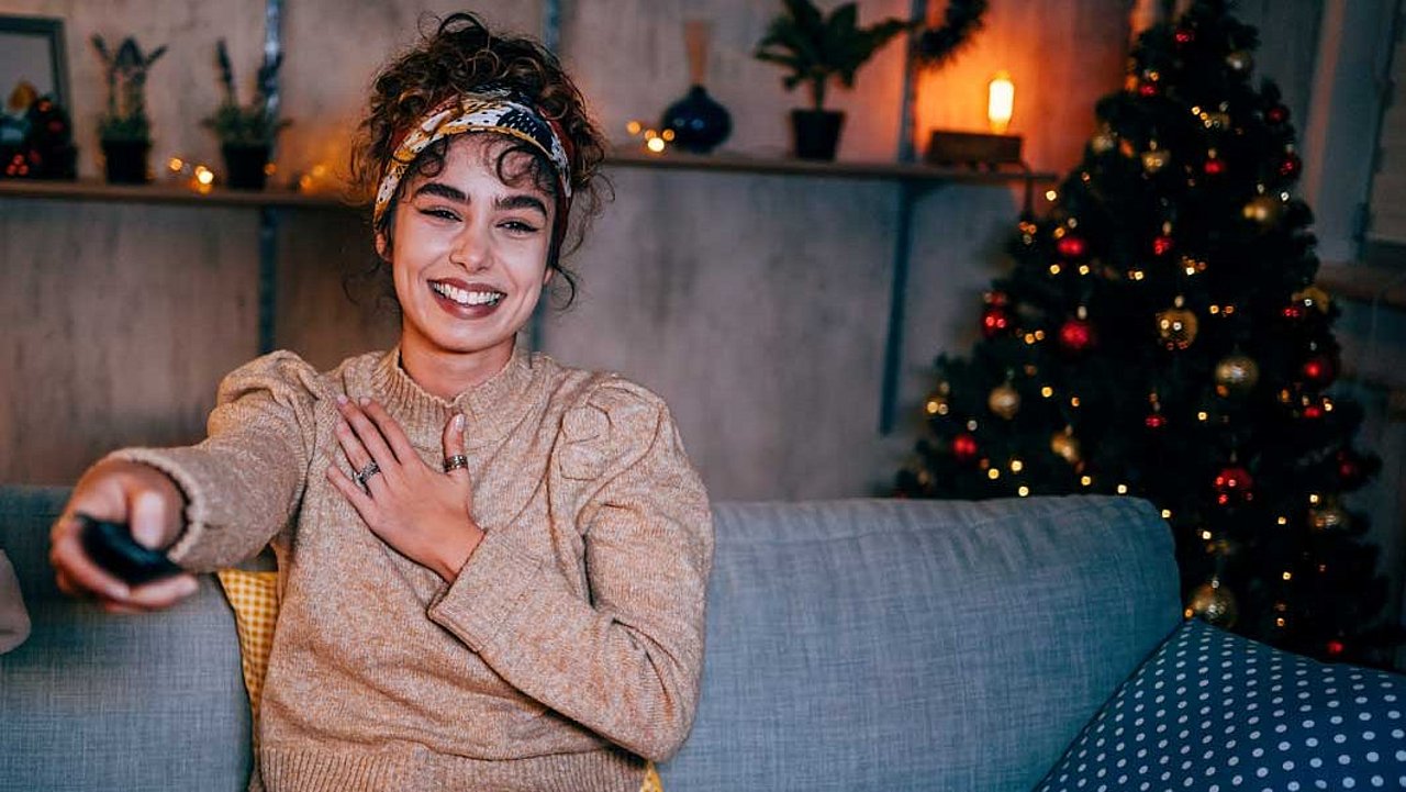 Frau auf Couch mit Fernbedienung in der Hand. Im Hintergrund ein Weihnachtsbaum und Weihnachtsdeko.