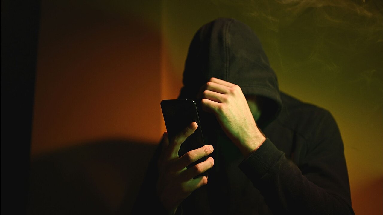 Eine anonyme Person in einem dunklen Raum, ein schwarzer Kapuzenpulli, die Kapuze tief ins Gesicht gezogen, den Kopf voller Scham gesenkt, in der Hand ein schwarzes Smartphone