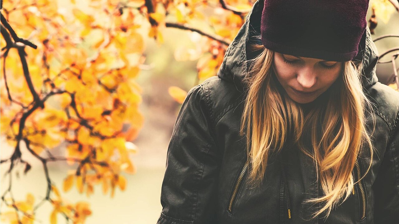 Eine Frau mit Mütze, langen blonden Haaren und grüner Jacke hält den Blick zu Boden gesenkt. Sie strahlt Melancholie und Nachdenklichkeit aus. Im Hintergrund buntes Herbstlaub und goldenes Licht