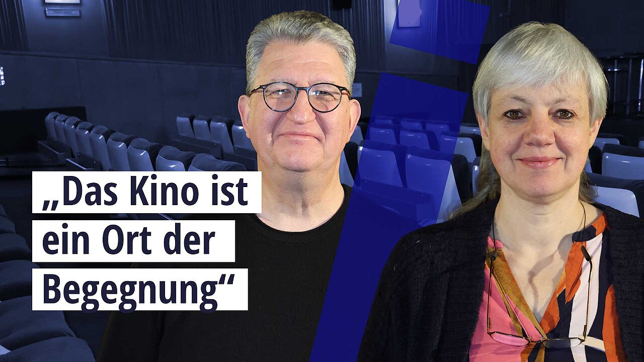 Zwei Personen, rechts eine Frau mit grauen Haaren, braunen Haaren und einer bunten Bluse, links ein Mann mit grauen Haaren, dunkler Brille und schwarzem Shirt. Beide stehen vor einem großen Kinosaal mit vielen leeren Sitzen. Text: "Das Kino ist ein Ort der Begegnung".
