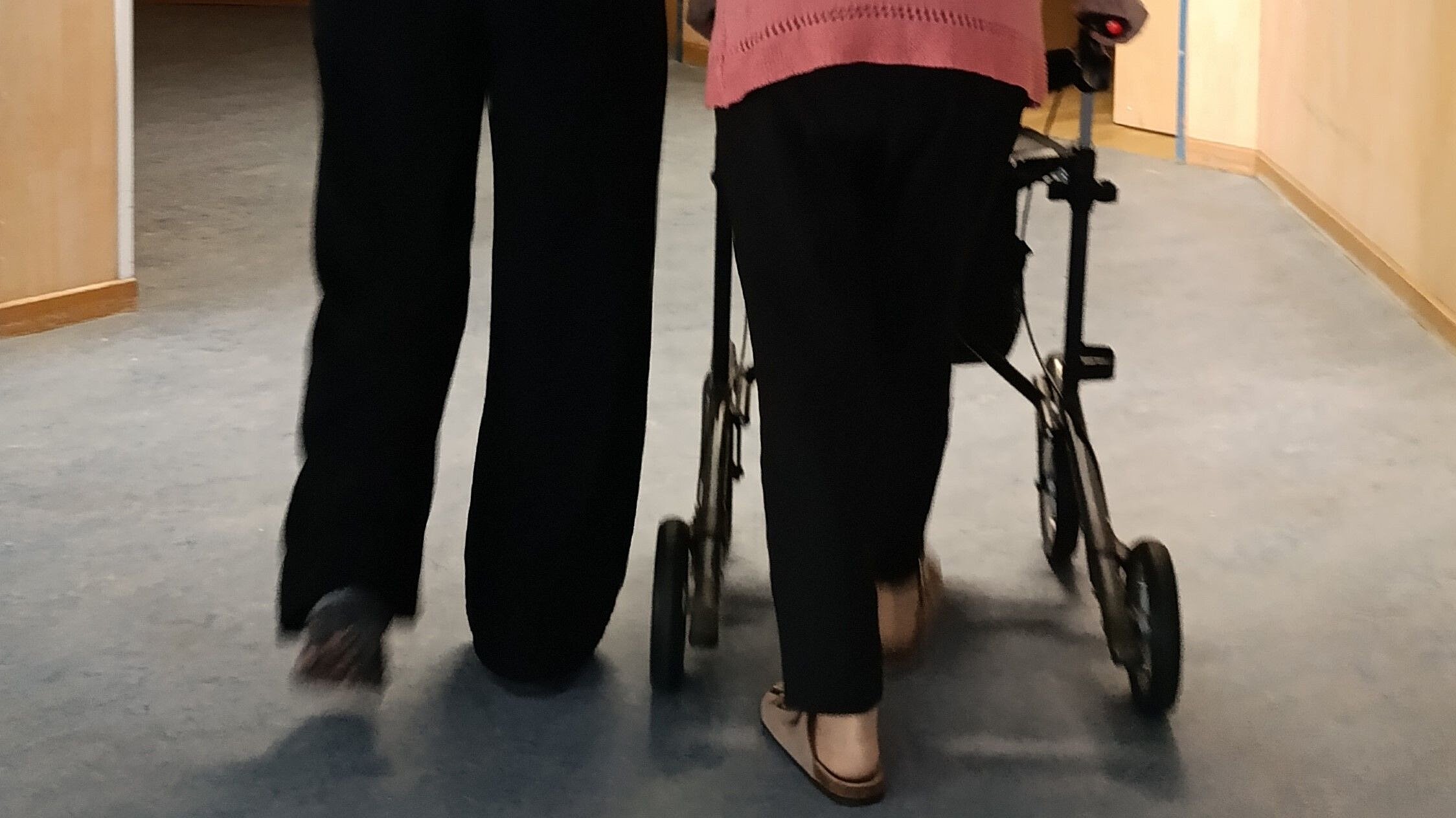 Zwei Personen gehen nebeneinander her. Nur von der Hüfte abwärts von hinten zu sehen. Rechts eine Person mit Rollator, links eine Pflegerin