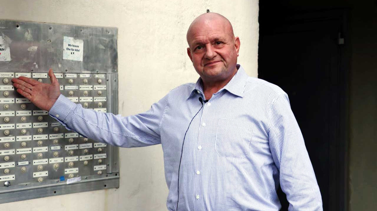 Achim Kaffenberger steht vor seiner Wohnung und zeigt auf seinen Namen auf dem Klingelschild