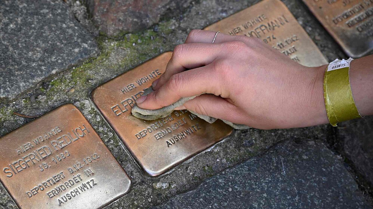 Eine Hand putzt mehrere Stolpersteine mit einem Lappen.