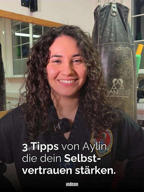 Eine Frau mit dunklen lockigen Haaren. Sie lächelt und trägt einen Taekwondo-Anzug. Das ist Aylin. Text: 3 Tipps von Aylin, die dein Selbstvertrauen stärken.
