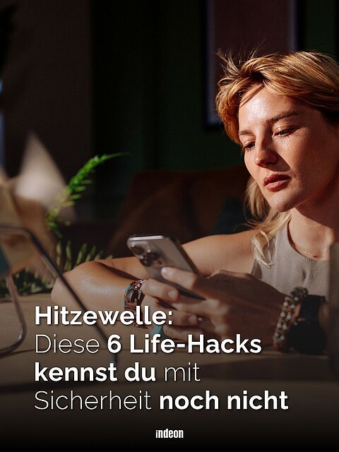 Eine Frau sitzt mit dem Smartphone in der Hand an einem Schreibtisch. ein kleiner Ventilartor vor ihr wirbelt Luft auf. 